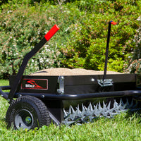 The Brinly Parts 40 Combination Aerator Spreader, model AS2-40BH-G, sits on green grass with its red-handled black frame filled with sand. It boasts spiked wheels for aeration and a 100 lb. capacity hopper for optimal soil penetration, all set against vibrant green foliage.