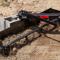 A Brinly Parts 38” Sleeve Hitch Box Scraper (BS-381BH) is used on a dry, uneven dirt surface for grading. Its attached to a concrete block with a partially visible label.