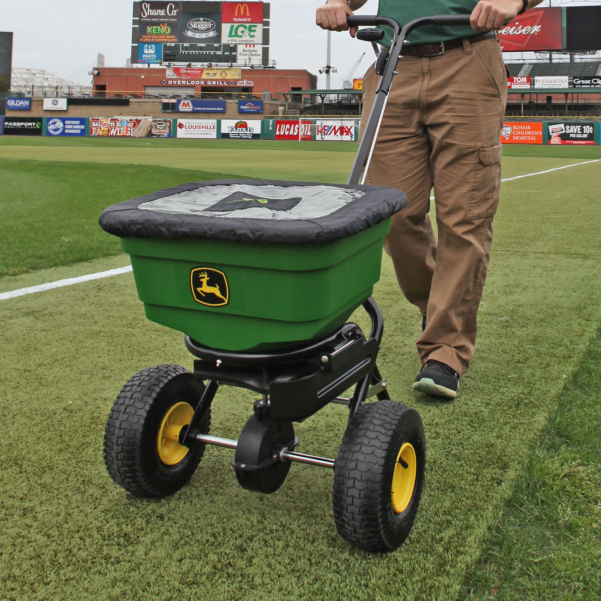 A person pushes a John Deere All-Season Push Broadcast Spreader (LP31340L-A) with an ergonomic split handle across a sports field, featuring robust black and yellow wheels, a durable poly hopper, and the iconic John Deere logo. The field is lined with advertisements in the background.