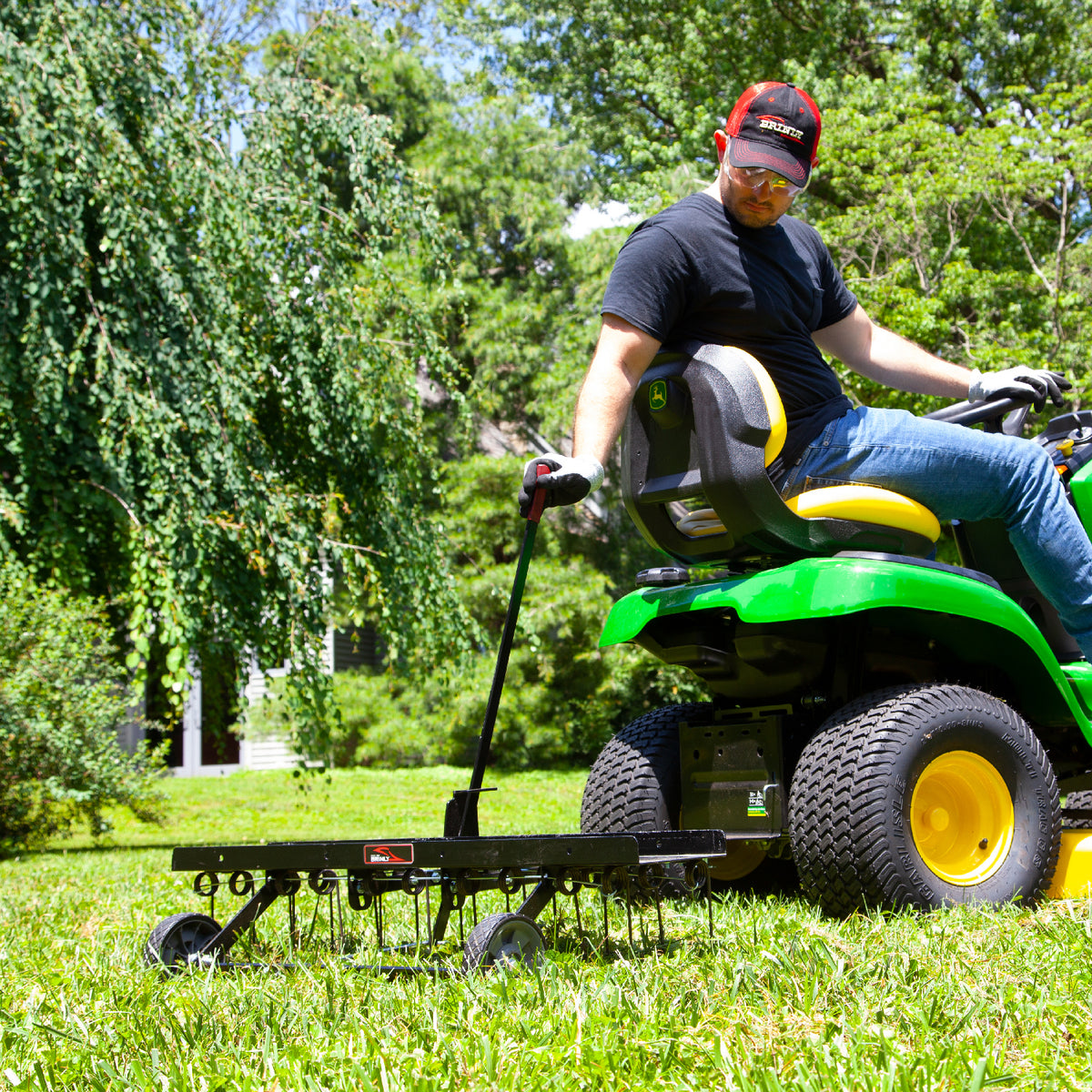 Wearing a red cap, black shirt, and jeans, a person skillfully operates a green lawn tractor with a Brinly Parts 40 Tow-Behind Dethatcher (DT2-40BH-G) on a sunny day. They remove thatch efficiently, surrounded by lush green trees and part of a house.