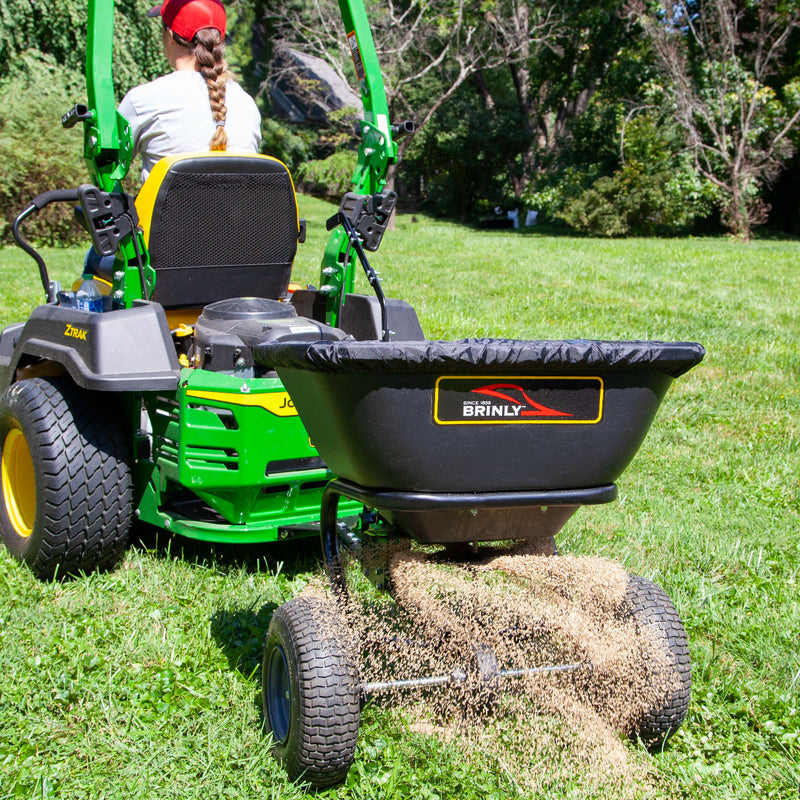 A person in a red cap drives a green lawn tractor, towing a Brinly Parts 125 LB. Tow-Behind Deluxe Spreader BS261BH-A with an extended handle and rust-proof polyethylene hopper across the grass, while trees and bushes provide a serene backdrop under the clear sky.