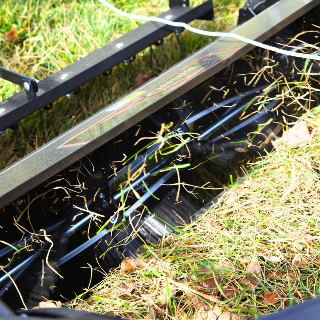 Close-up of the Brinly Parts 54" BrinlySweep DKMHP in action, its metal tines pulling up dead grass and leaves from a green lawn, with thatch and debris collected inside the machine.