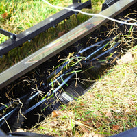 Close-up of the Brinly Parts 54" BrinlySweep DKMHP in action, its metal tines pulling up dead grass and leaves from a green lawn, with thatch and debris collected inside the machine.
