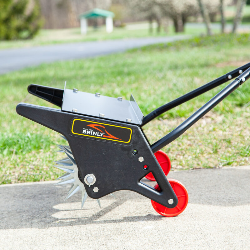 A Brinly Parts 18 Push Spike Aerator with 3D galvanized steel tines and red wheels stands on concrete, showcasing its spiky metal stars for effective lawn aeration. Grass and a blurred outdoor background complete the scene.