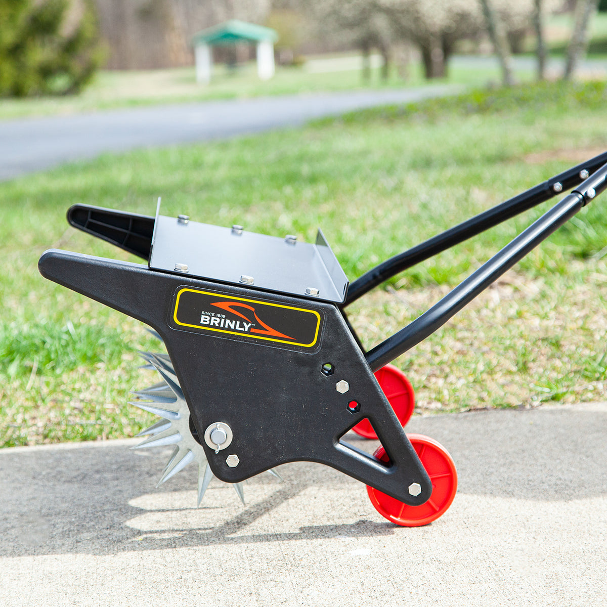 A side view displays the 18 Push Spike Aerator by Brinly Parts with spiked wheels, a metal frame of galvanized steel, and bright red plastic wheels. It rests on concrete near grass, showcasing the Brinly logo amidst trees and a blurred lawn aeration backdrop.