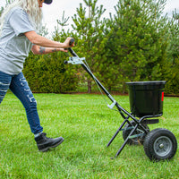 Wearing a gray shirt, jeans, and boots, a person uses the Brinly Parts 50 lb. Push Spreader | P20-500BH on green grass with evergreen trees in the background. The spreaders large handle and wheels ensure even distribution of lawn fertilizer across the yard.