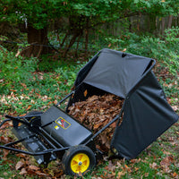The Brinly Parts 42″ Lawn Sweeper with Dethatcher (STS-42BHDK-A) collects fallen leaves in a grassy, tree-surrounded yard, its yellow-wheeled container partially filled with dry leaves.