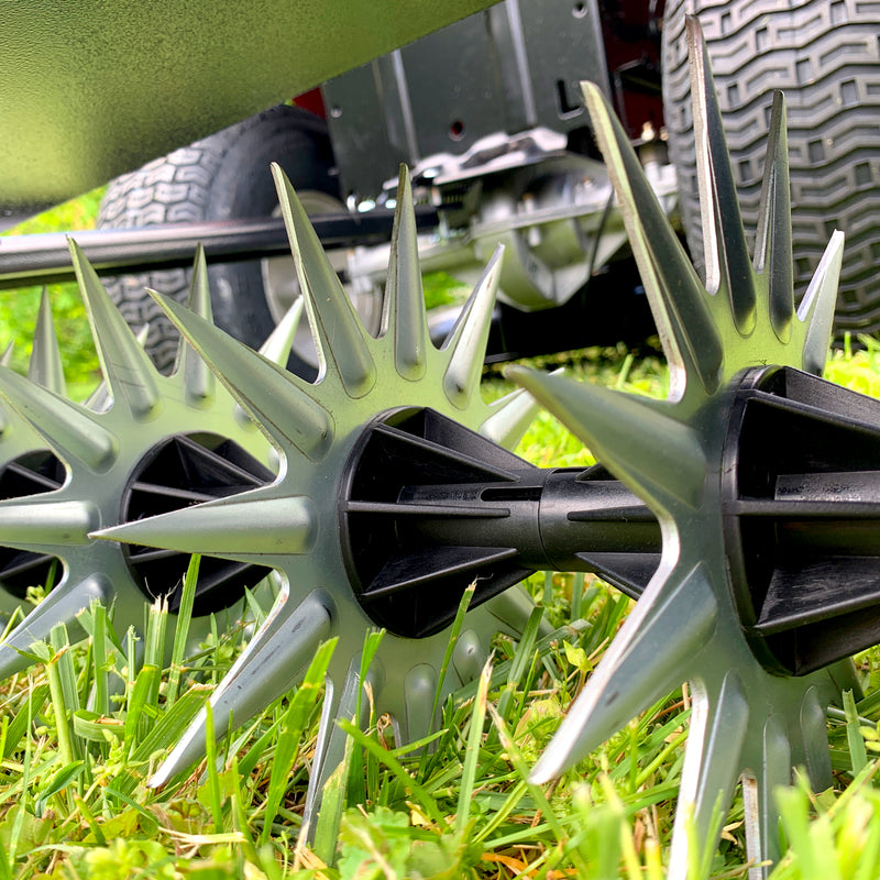 The Brinly Parts 40″ Tow-Behind Spike Aerator (SAT2-40BH-G) features spiked metal wheels on lush green grass, designed for efficient soil aeration. Its set against large tires and a partially visible machine body with its wide channel tow bar & transport wheels.