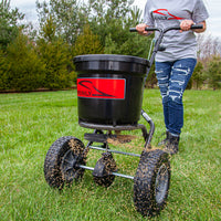 A person in ripped jeans and a gray shirt pushes the Brinly Parts 50 lb. Push Spreader | P20-500BH, with its large wheels and red label, efficiently distributing fertilizer across the grass as tall trees line the background.