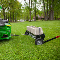 A Brinly Parts 40″ Spike Aerator with Transport Wheels & 3D Galvanized Steel Tines (SAT-401BH) is towed by a green mower with a black cart carrying concrete blocks, secured by a strap, on a grassy lawn surrounded by trees and houses.