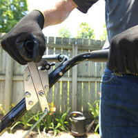 A person in black gloves adjusts the clutch cable on a riding lawn mowers steering wheel. Nearby, a Brinly Parts 50 lb. Push Spreader (P20-500BH), a kettle grill, wooden fence, and lush plants are visible as they work outdoors with a trusty wrench.