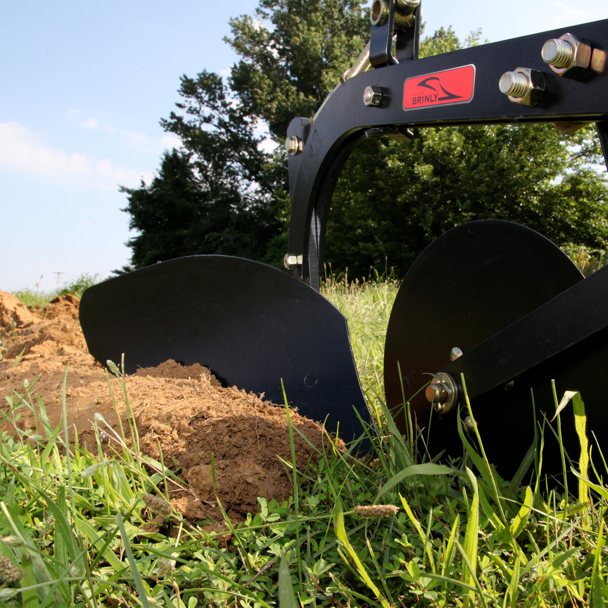 Close-up of a black Brinly Parts Sleeve Hitch Moldboard Plow | PP-510s dual blades cutting through earth surrounded by green grass, a pile of soil in front, with trees and blue sky in the background.