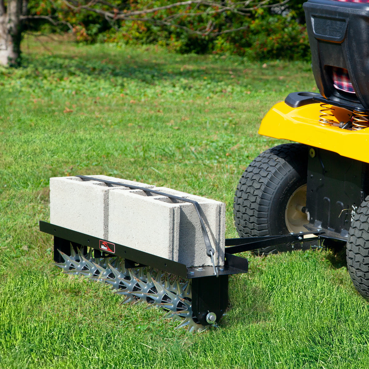 A yellow lawn tractor tows a Brinly Parts Spike Aer 40 spike aerator, loaded with three concrete blocks, across a grassy lawn with trees in the background.