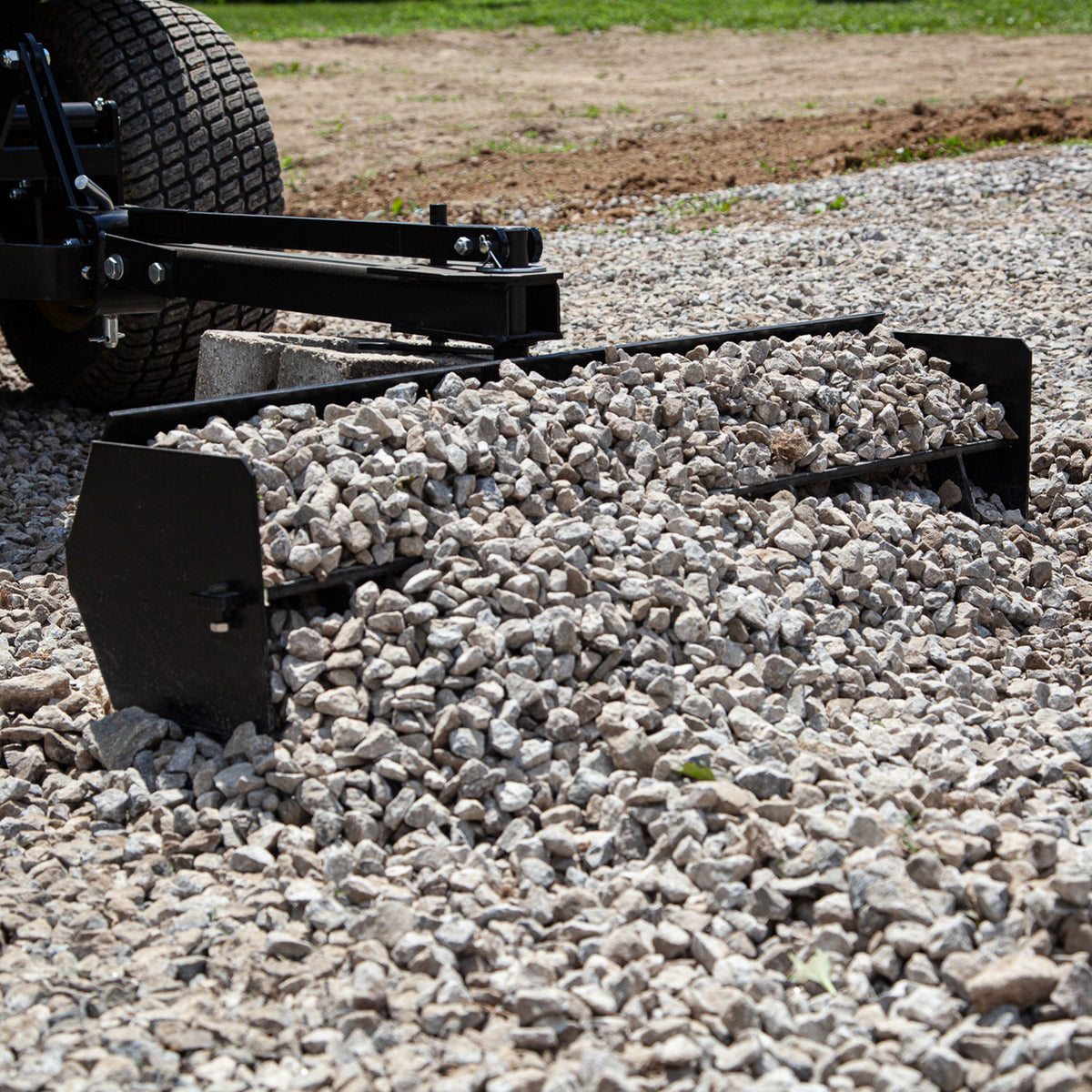 A 38” Sleeve Hitch Box Scraper, model BS-381BH by Brinly Parts, expertly grades a gravel driveway, spreading and smoothing an uneven pile of small gray stones with its rear blade attachment.