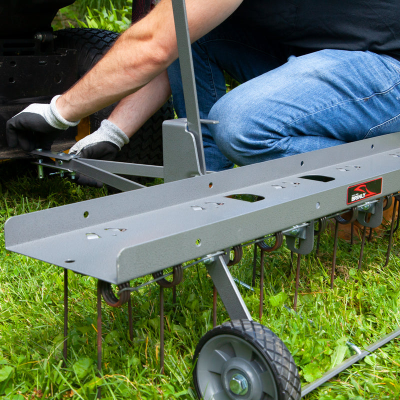 A person wearing gloves attaches a Brinly Parts 40″ Dethatcher in Hammered Gunmetal (DT2-40BH-S) to a tractor. The focus is on the tow-behind dethatcher with tines and wheels, resting on green grass.