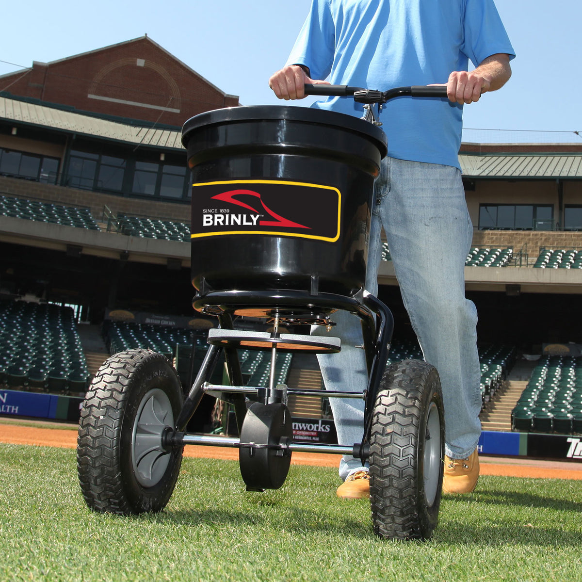 A person in jeans and tan shoes uses a Brinly Parts 50 LB. Push Spreader with Side Deflector & Hopper Cover (P20-500BHDF-A2) on a sports field, with stadium seats and buildings seen in the background.