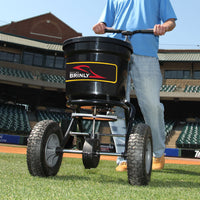 A person in jeans and tan shoes uses a Brinly Parts 50 LB. Push Spreader with Side Deflector & Hopper Cover (P20-500BHDF-A2) on a sports field, with stadium seats and buildings seen in the background.