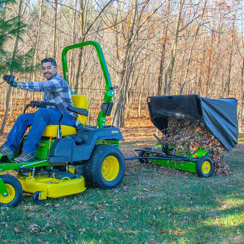 A man in a striped sweater operates a green and yellow ride-on lawn mower, using a John Deere 42 in. 24 cu. ft. Tow-Behind Lawn Sweeper filled with autumn leaves. He smiles while working on grassy terrain with sparse foliage trees in the background.