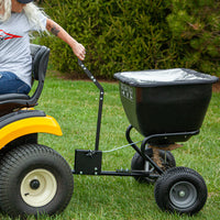 A person rides a yellow lawn mower towing a Brinly Parts 175 LB. Tow-Behind Deluxe Spreader, model BS361BH-A, half-filled with seed or fertilizer in its rust-proof polyethylene hopper, attached via a universal hitch. Grass and trees stretch in the background.