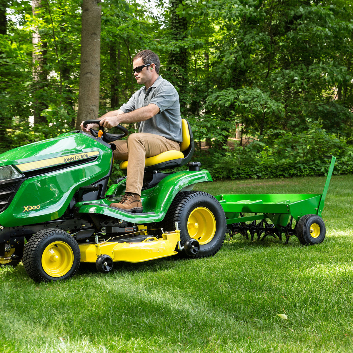 A person drives a John Deere X300 mower towing a small green trailer across lush lawn, equipped with a Brinly Parts Plug Aerator - 48 in Tow-Behind, set against dense foliage. Carburized steel tines promise to rejuvenate the grass.