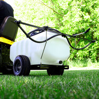 The Brinly Parts 25 Gallon Tow-Behind Sprayer | ST-251BH with Ultra Lo-Drift™ Spray Tips is attached to a tractor on a grassy area. It features a white tank, two black wheels, and stands ready under the sun with lush green trees enhancing the serene landscape.