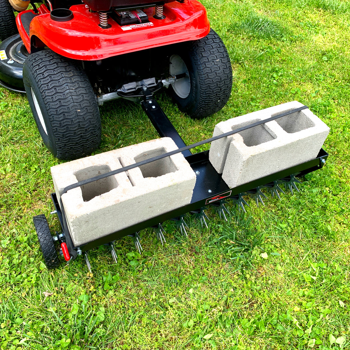 A red lawn tractor pulls a Brinly Parts 40″ Tow-Behind Spike Aerator, model SAT2-40BH-G, on grass. The black aerators wide channel tow bar and two large concrete blocks enhance its efficiency in removing thatch and improving lawn aeration.