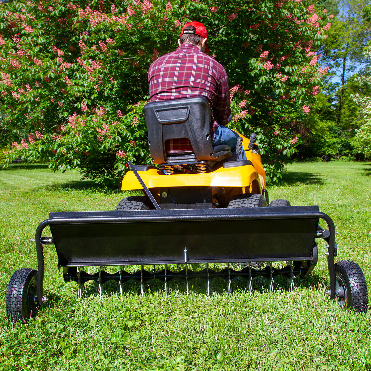 A person wearing a red plaid shirt and cap operates a yellow lawn tractor while towing the Brinly Parts 40 Combination Aerator Spreader with Pneumatic Tires (AS2-40BH-G) featuring a 100 lb. capacity across a green lawn, with a blooming bush in the background under the clear blue sky.