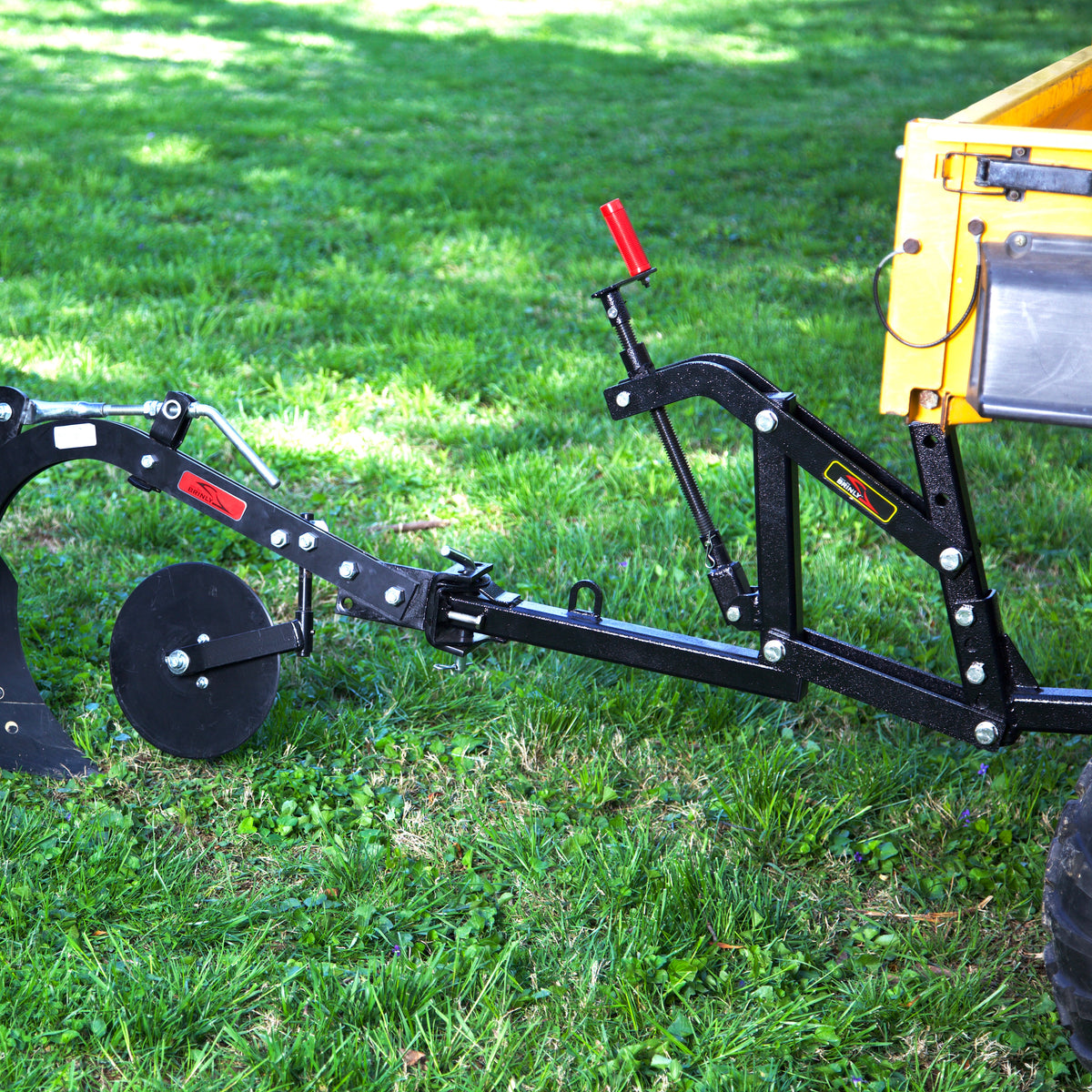 A close-up of the Brinly Parts Universal ATV/UTV One-Point Lift (OPL2-BH) attached to a black plow on a yellow tractor, set on green grass with sunlight streaming through trees in the background.