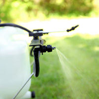 The Brinly Parts ST-251BH Tow-Behind Sprayer features Ultra Lo-Drift™ spray tips, releasing a fine mist of liquid fertilizer. The sprayer is attached to a 25-gallon plastic tank with hoses, set against a backdrop of blurred grass and foliage.