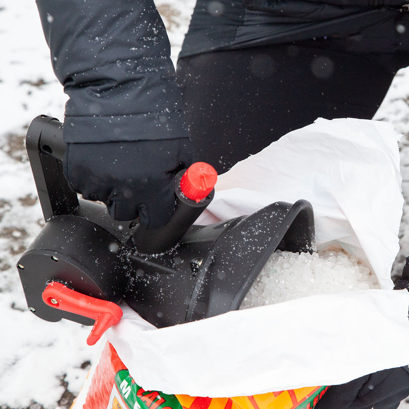 A person in black gloves uses a Brinly Parts 5lb Capacity Handheld Spreader | HHS3-5BH, featuring a red handle, to distribute ice melt pellets from a bag onto the snowy ground as snowflakes gently fall and some pellets are visible in the air.