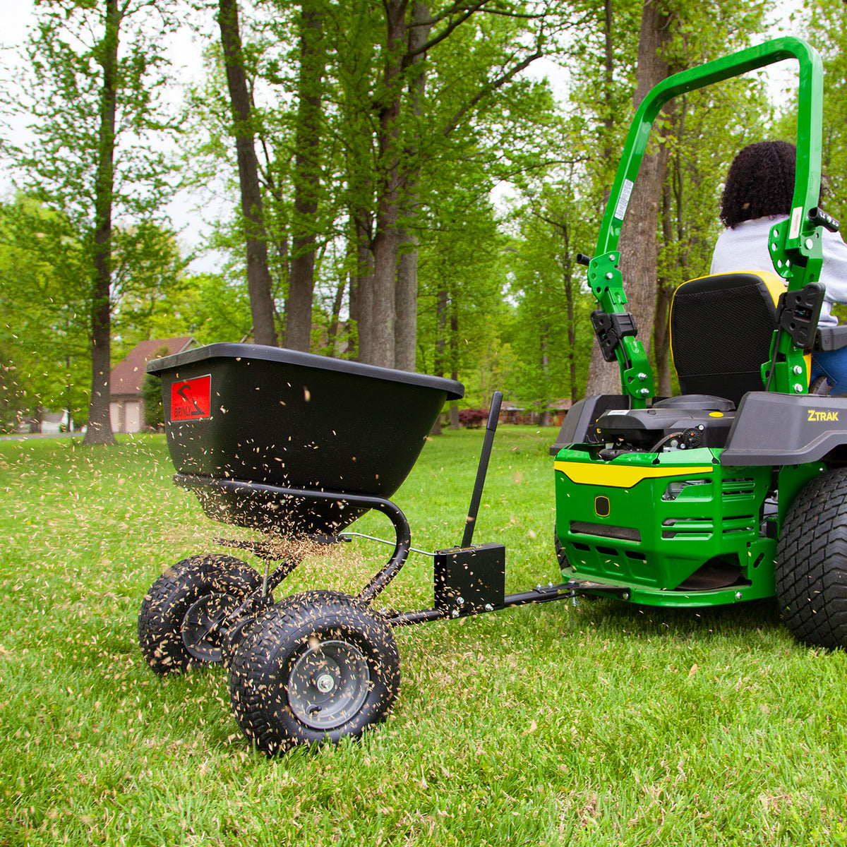 A person uses a Brinly Parts 125 Lb. Tow-Behind Broadcast Spreader (BS26BH) with a rust-proof polyethylene hopper to evenly distribute seeds across a green lawn, creating an effective fertilization. Tall trees and a house in the background suggest a serene spring setting.