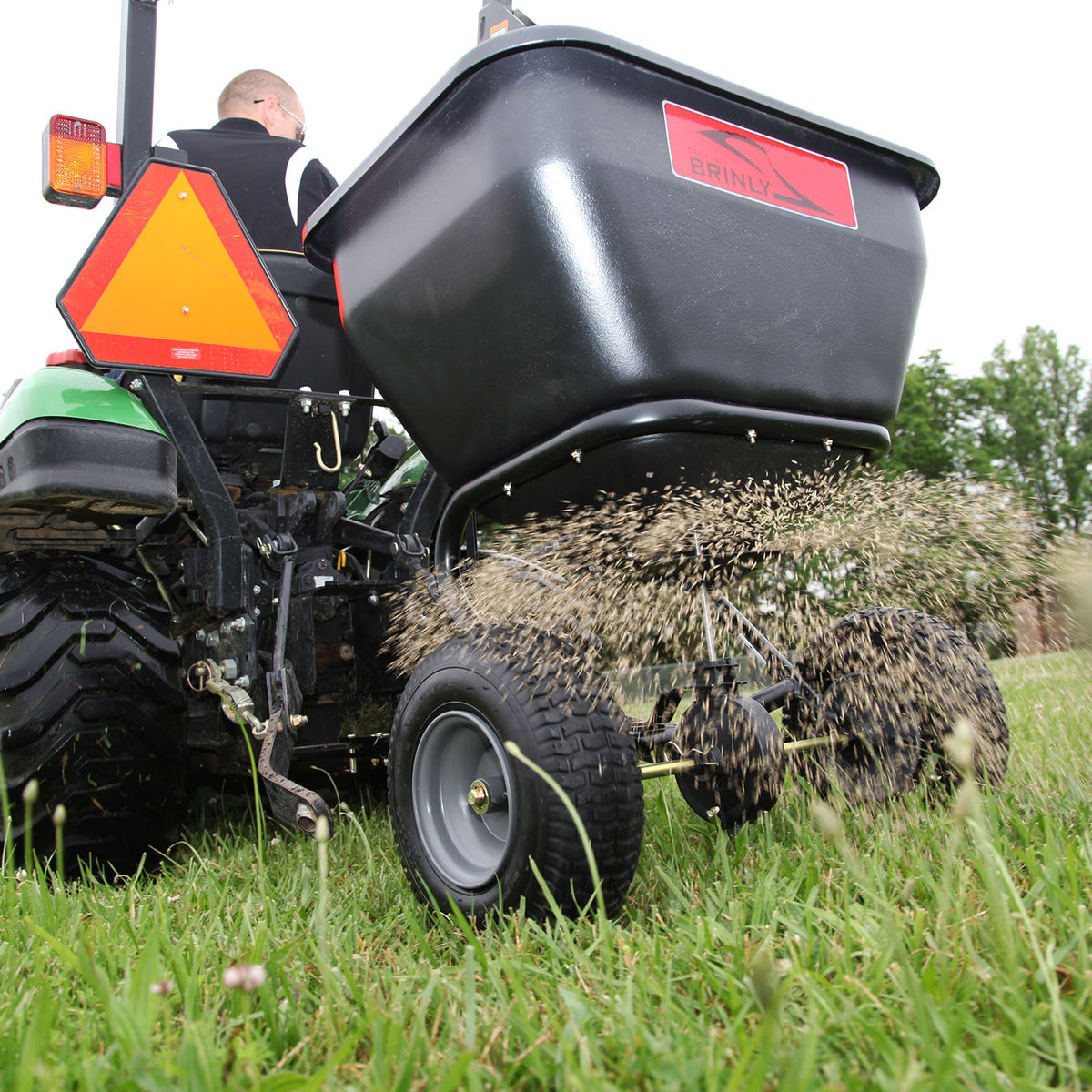 Using the rust-proof 175 lb. BS36BH Tow-Behind Spreader from Brinly Parts, a person disperses seeds efficiently across a grassy lawn attached to a tractor. The black spreader with its red label performs well among foreground weeds, with tall trees in the background.