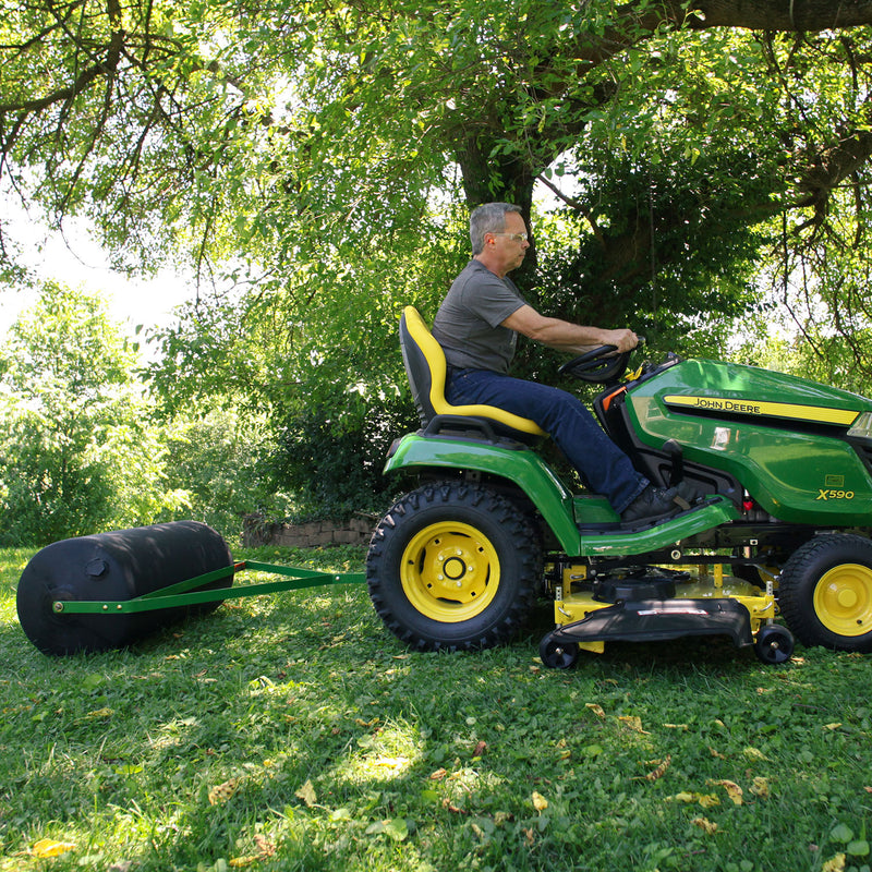 A man is mowing grass on a green riding lawn mower, towing a John Deere 36 Lawn Roller (PRT-361SJD/LPPRT36JD) with a tethered hitch pin. Under the large leafy tree, dappled sunlight creates a serene and natural backdrop.