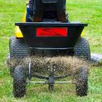 A person on a yellow lawn tractor uses the Brinly Parts BS26BH 125 Lb. Tow-Behind Broadcast Spreader to evenly distribute grass seed on the lawn. The rust-proof polyethylene hopper ensures longevity as seeds visibly scatter, ensuring an even blanket of new growth.