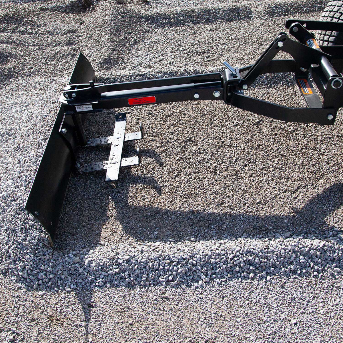 The Brinly Parts 42” Sleeve Hitch Box Scraper (BS-42BH) attached to a tractor efficiently levels a gravel driveway, utilizing its sturdy carbon steel blade for even distribution and smooth surface results.