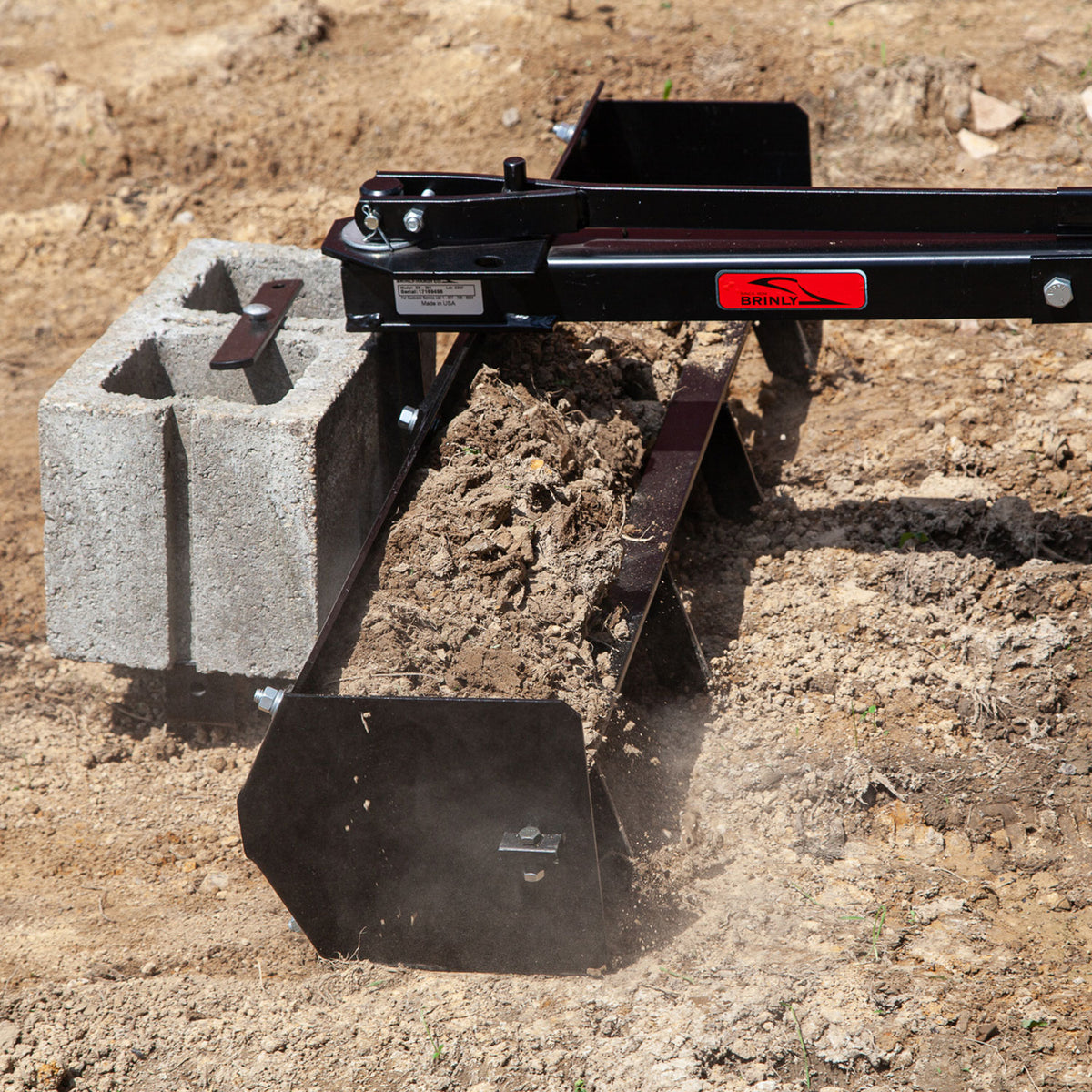 A close-up shows the Brinly Parts 38” Sleeve Hitch Box Scraper (BS-381BH), with a steel frame and added concrete blocks for weight, paired with a rear blade. Its moving on dry, dusty soil, creating a small dust cloud, indicating its preparing the ground for planting or grading.