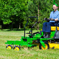 Wearing denim, gloves, and a green headband, a person skillfully steers a John Deere Front-Mount 48 ZTR Dethatcher (CPLP83252) with its yellow seat and black wheel across the grassy area surrounded by trees.