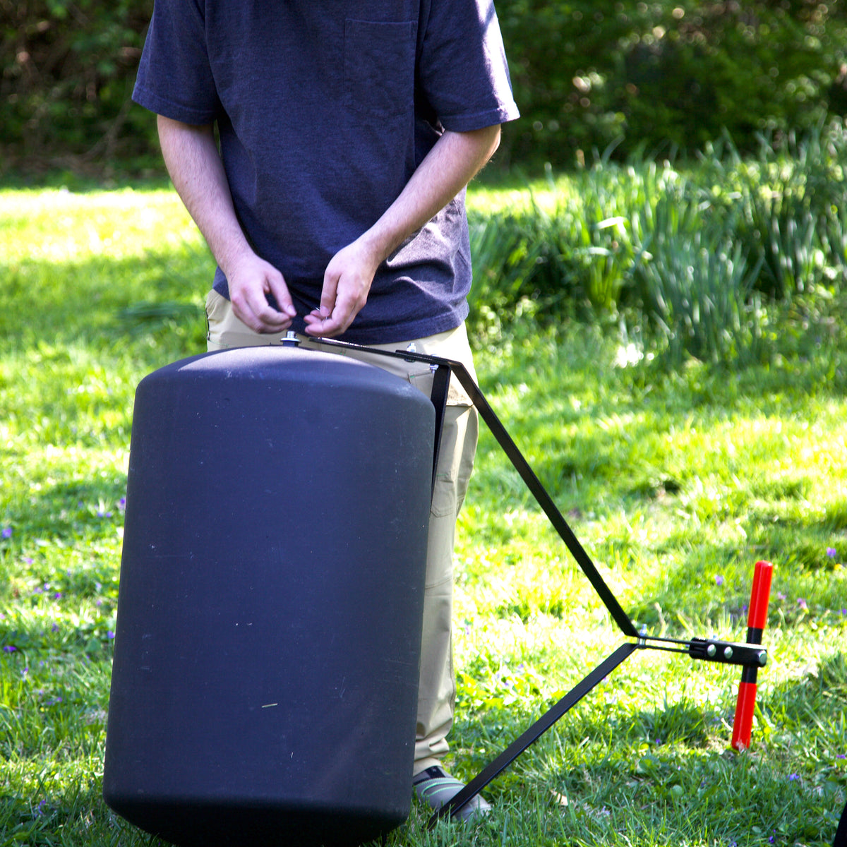 A person stands on grass, adjusting a Brinly Parts 28 Gallon Push or Tow Poly Lawn Roller (PRC-243BH) with a red handle, surrounded by greenery and sunlight. The person's face is not visible.