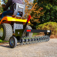 A person drives a yellow lawn tractor pulling the Brinly Parts 40″ Spike Aerator with Transport Wheels & 3D Galvanized Steel Tines (SAT-401BH) across a concrete surface in a garden with trees and shrubs in the background.