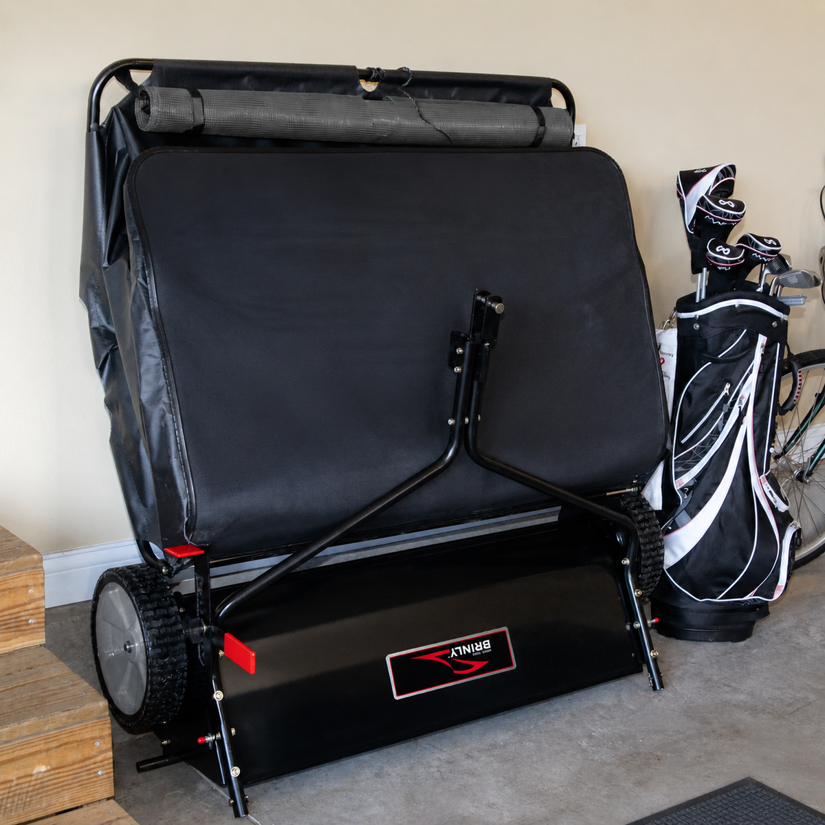 A Brinly Parts 42″ Lawn Sweeper with DOUBLE-HELIX Brushes (model LS2-42BH2-G), featuring a large black body with red accents, is parked in a garage next to a set of golf bags and clubs. Wooden steps are visible on the left, and part of a bicycle appears on the right.