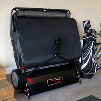 A Brinly Parts 42″ Lawn Sweeper with DOUBLE-HELIX Brushes (model LS2-42BH2-G), featuring a large black body with red accents, is parked in a garage next to a set of golf bags and clubs. Wooden steps are visible on the left, and part of a bicycle appears on the right.