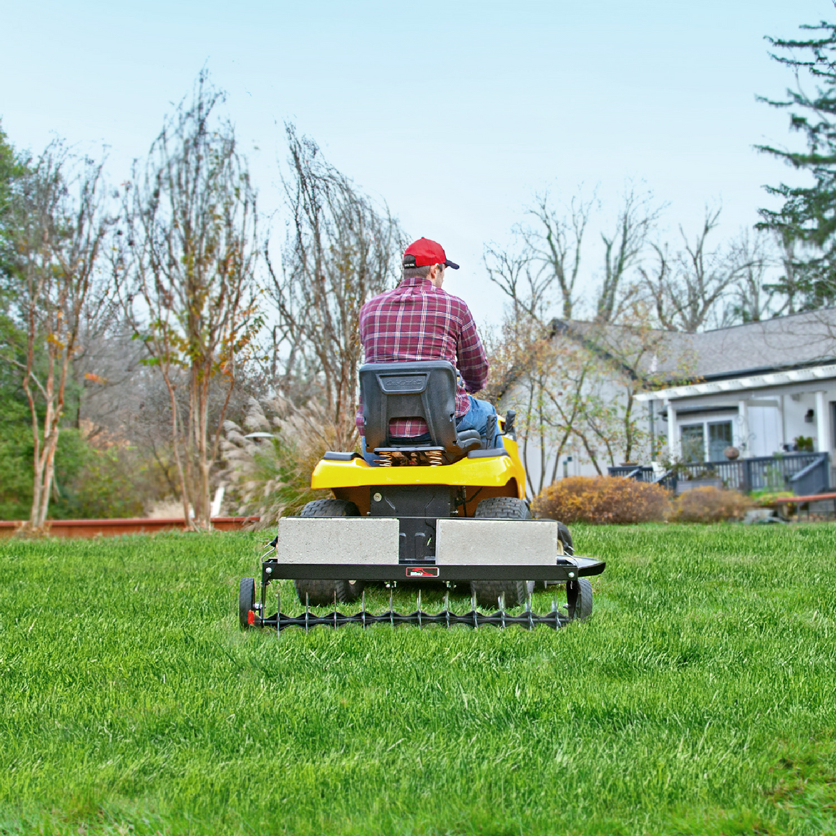 A person in a red cap and plaid shirt uses a yellow lawn tractor to pull the Brinly Parts 40" Tow-Behind Spike Aerator (SAT2-40BH-P2) with double tow bar and transport wheels across a green yard, house and bare trees behind.