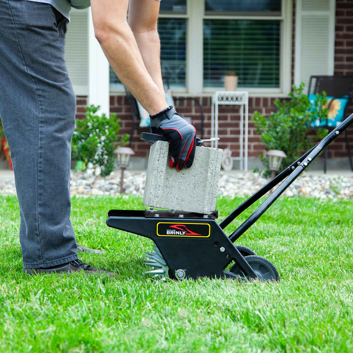 Wearing gloves, someone places a concrete block onto a Brinly Parts 20 Push Spike Aerator with 3D Galvanized Steel Tines on a grassy lawn to improve soil aeration in front of a brick house with a porch and plants.