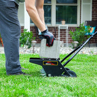 Wearing gloves, someone places a concrete block onto a Brinly Parts 20 Push Spike Aerator with 3D Galvanized Steel Tines on a grassy lawn to improve soil aeration in front of a brick house with a porch and plants.