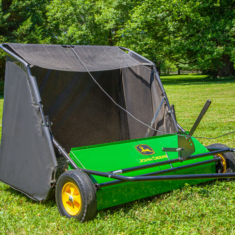 A John Deere 42 in. 24 cu. ft. Tow-Behind Lawn Sweeper with yellow wheels is on a grassy lawn under green trees and a blue sky, featuring a large black collection bag and sturdy metal frame.