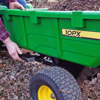 In a garden, someone tilts the lever of a John Deere 180 Degree Full Dump Cart, featuring a 650 lb. capacity and an 180-degree dump angle. The green 10PX tow-behind cart, filled with dark mulch, sits on black wheels with yellow hubs among dry leaves.