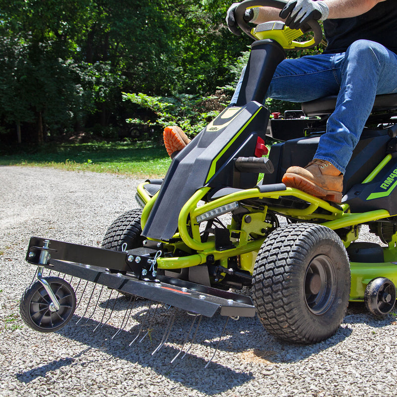 A person in jeans and brown boots operates a RYOBI Electric Riding Mower on gravel, using the Brinly Parts 38 in. Front Mount Dethatcher (DT-38RY), featuring tines and a wheel for precision landscaping, with tall trees in the background completing this scene of expert lawn care.