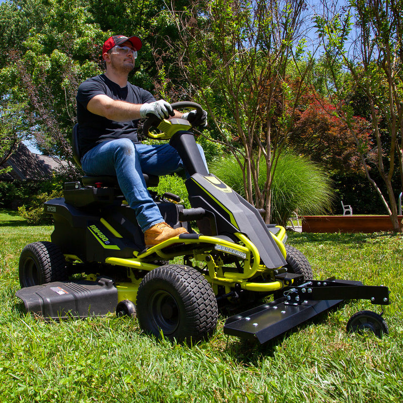 A person in a red cap, sunglasses, gloves, black shirt, and jeans is riding a RYOBI Electric Riding Mower with a 38 in. Front Mount Dethatcher by Brinly Parts across a grassy lawn. Trees and a planter are in the background under clear blue skies, highlighting their dedication to lawn care.