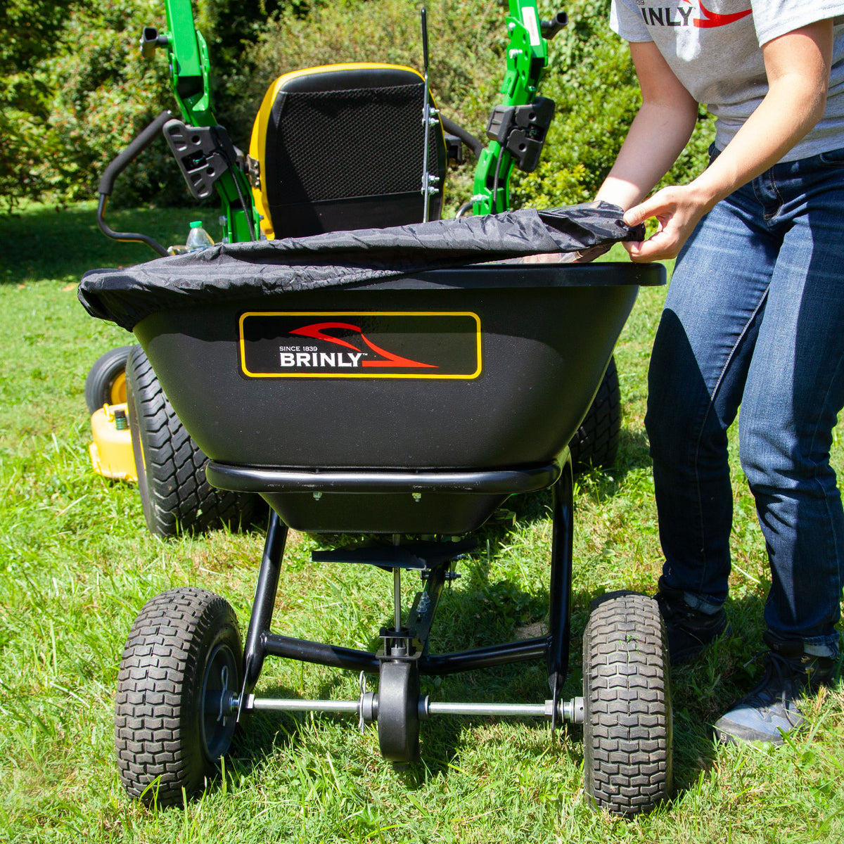 A person operates a Brinly Parts 125 LB. Tow-Behind Deluxe Spreader with Extended Handle & Cover, model BS261BH-A, attached to a green and yellow tractor on grass. The open rust-proof polyethylene hopper is being adjusted under the bright sun, creating an ideal scene for lawn fertilization.