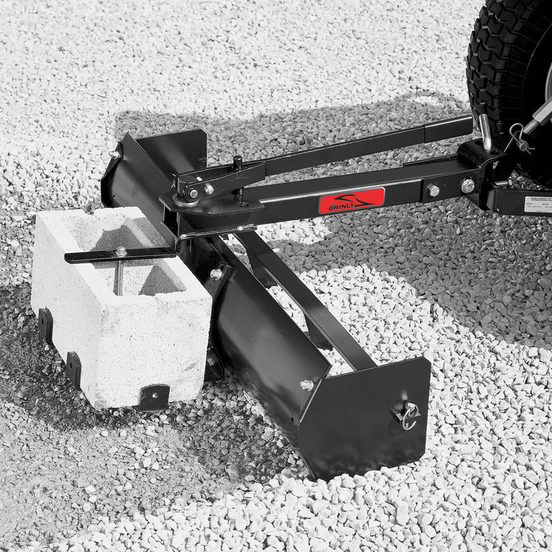 The Brinly Parts 38” Sleeve Hitch Box Scraper (BS-381BH), featuring a red label, is attached to a wheeled machine on gravel with a concrete cinder block for additional grading weight.